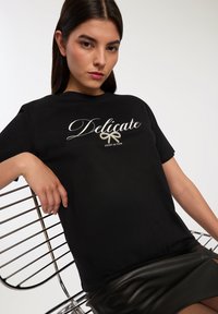 Czarna bawełniana koszulka z napisem "Delikatny" w białym kolorze, otoczonym grafiką wstążki. Nosi ją modelka w czarnej skórzanej spódnicy. Siedzi na metalowym krześle.