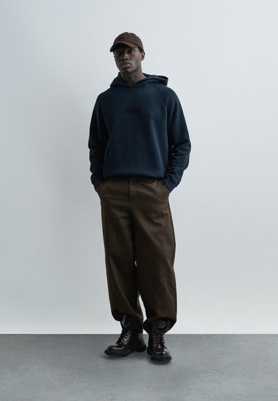 Άνδρας με σκουρόχρωμο μπλε φούτερ με κουκούλα, καφέ παντελόνι φαρδιάς γραμμής, καφέ δερμάτινες μπότες και καφέ καπέλο, στέκεται μπροστά σε ένα απλό ανοιχτό φόντο.