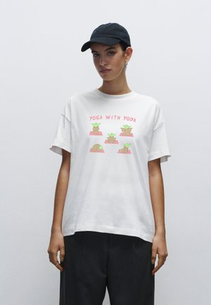 Witte katoenen T-shirt met een cartoonprint van vier Yoda-figuren die yoga poses doen, met de tekst "Yoga Met Yoda" in het roze.