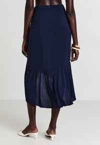 Donna che indossa una gonna midi blu navy con orlo a balze, sandali con tacco bianchi e bracciali crema e marrone, vista da dietro.