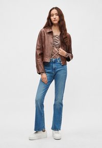 Chaqueta corta de cuero marrón sobre una blusa con estampado, combinada con vaqueros acampanados azules de cintura alta y zapatillas blancas. Diseño sencillo, estilo casual.