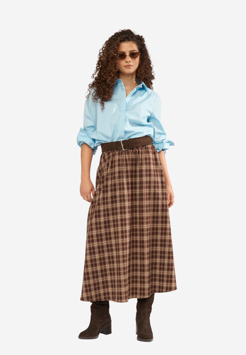 Femme aux cheveux bouclés portant des lunettes de soleil, une chemise bleu clair, une jupe midi à carreaux marron avec ceinture et des bottes marron foncé, debout devant un fond blanc.