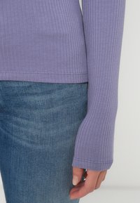 Langermet ribbestrikket topp i lys lilla, laget av myk stoff, sammen med blå denimbukser. Ermeløsningen har et tettsittende mansjett-design.