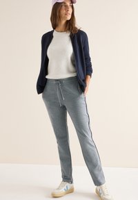 Lichtgrijze sportbroek met zijstrepen, gecombineerd met een marineblauwe bomberjack en een lichte trui, accentueel met een roze pet en witte sneakers.