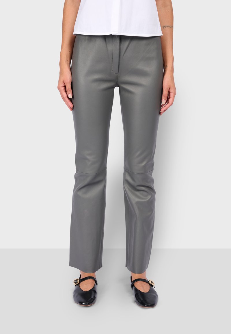 Vrouw die grijze leren broek en zwarte Mary Jane schoenen draagt, staand tegen een effen lichtgrijze achtergrond.