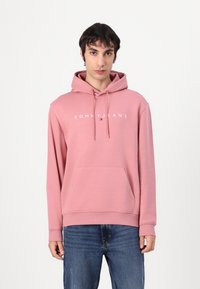 Hoodie rosa chiaro con coulisse regolabile, tasca frontale e logo bianco "TOMMY JEANS". Abbinato a jeans blu, mostrato su uno sfondo semplice.