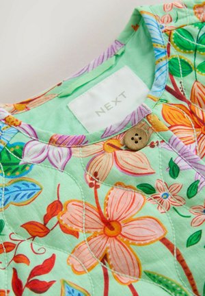 Zelená látka s květinovým vzorem růžových, oranžových a modrých květin. Má kulatý výstřih a dřevěné knoflíkové zdobení u límce.