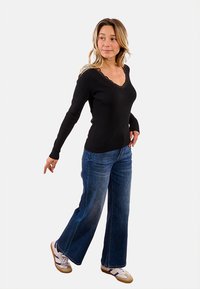 Donna con un top nero a maniche lunghe e jeans blu a gamba larga, mentre cammina e guarda oltre la spalla, indossando sneakers bianche con strisce viola.