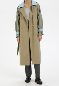 Person klädd i en beige trenchcoat med blå krage och manschetter, grå randiga byxor, grå topp och svarta loafers som står mot vit bakgrund.