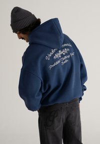Bărbat îmbrăcat cu un hanorac bleumarin pe spate cu broderia "Verdant Reverie Prohibited Clothing Berlin" și blugi negri cu logo brodat pe buzunar.