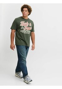 Groen T-shirt met korte mouwen met witte en rode tekst, gecombineerd met blauwe jeans en grijze sneakers. Model loopt tegen een effen achtergrond.