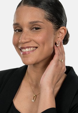 Gold-tone jewelry features a geometric earring and a pendant necklace with a crystal accent, worn by a model in a black top.