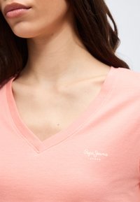 Γυναίκα με απαλό ροζ μπλουζάκι V με μικρό λευκό λογότυπο "Pepe Jeans London" στο αριστερό στήθος, που εμφανίζει τον λαιμό και την άνω περιοχή του στήθους.