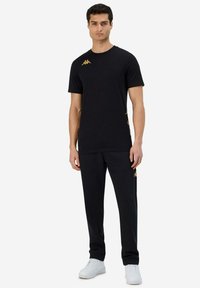 Zwarte t-shirt met een gouden logo, gecombineerd met zwarte joggingsbroek. Schoenen zijn wit. Gladde textuur, aansluitend ontwerp. Accenten langs de zijkant.