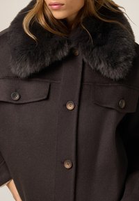 Cappotto in lana marrone scuro con colletto in pelliccia sintetica, grandi tasche a toppa e chiusure con bottoni. Tessuto morbido con una silhouette strutturata.