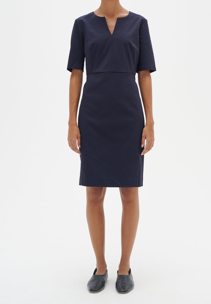 Femme portant une robe bleu marine foncé longueur genou avec manches courtes et décolleté en V, associée à des chaussures plates noires sans lacets.
