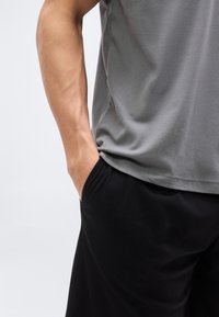 Άτομο που φοράει γκρι πουκάμισο και μαύρο παντελόνι με το ένα χέρι χαλαρά μέσα στην τσέπη του παντελονιού, μπροστά σε απλό φόντο.