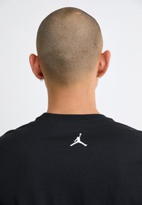 La maglietta nera in cotone presenta un logo Jumpman bianco nella parte superiore della schiena. La maglietta ha un classico collo girocollo e maniche corte.