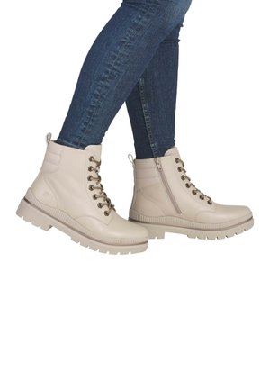 Beige Schnür-Stiefeletten mit strukturierter Oberfläche, seitlichem Reißverschluss, Gummisohle und metallischen Ösen, kombiniert mit eng anliegenden dunklen Jeans.