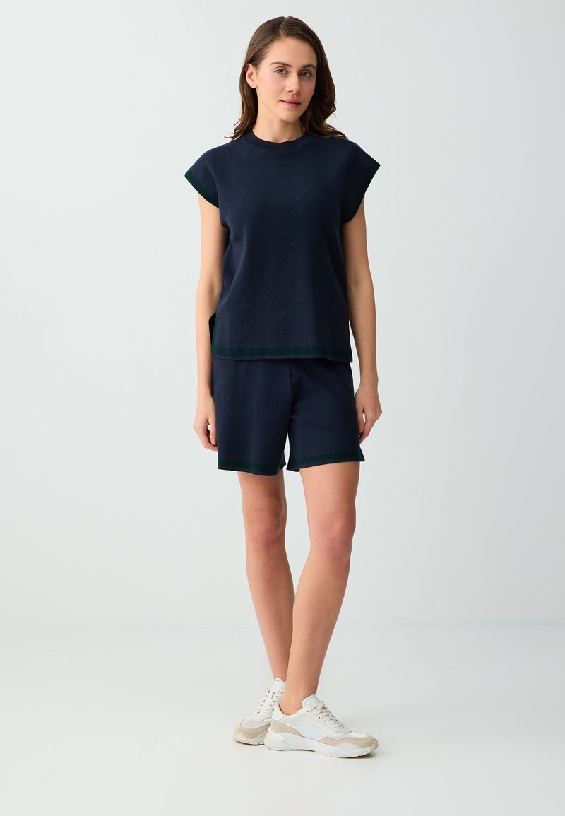 Femme debout portant un haut sans manches bleu marine et un short assorti avec des baskets blanches sur un fond uni gris clair.