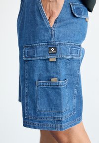 Sorturi din denim de culoare albastră medie, cu multiple buzunare, un eticheta neagră Converse și o croială lejeră cu detalii vizibile de cusătură.