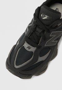 Fekete sportcipő hálós és szintetikus részletekkel, ívelt alakzatú, jól látható logóval és tapadásos texturált talppal.