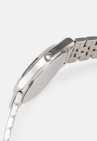 Montre en acier inoxydable argenté avec un boîtier rond, couronne texturée et bracelet en mailles. La montre présente une finition métallique lisse et des lignes épurées.