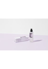Bouteille en verre transparent avec un couvercle de pompe noir, contenant un produit liquide. Boîte blanche avec des accents violets et du texte. Fond violet doux.