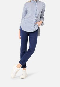 Osoba ubrana w niebieską i białą pionową w paski koszulę z guzikami, granatowe spodenki z mankietami i białe trampki, stoi z jedną ręką w kieszeni.