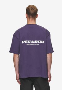 Violeta pārāk liela t-kreklu ar baltu "PEGADOR" logotipu un tekstu "SERTIFICĒTS AUTENTISKS IELA MODES APĢĒRBS" uz muguras.
