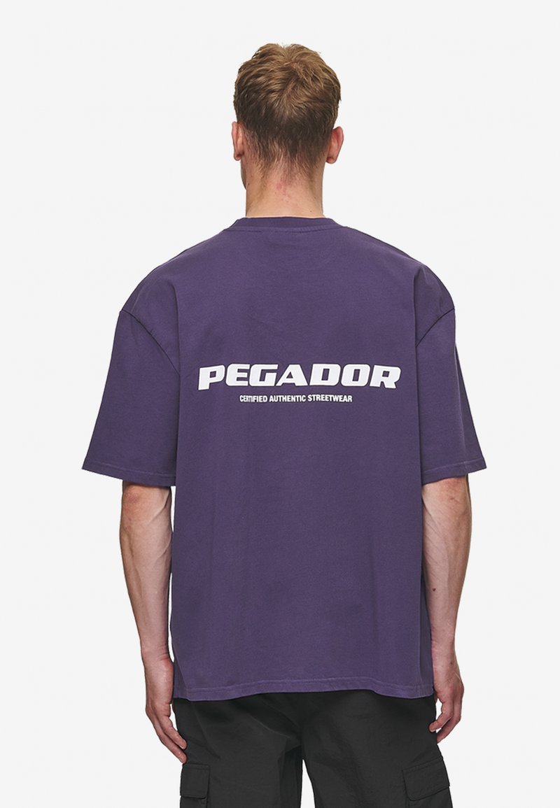 Violeta pārāk liela t-kreklu ar baltu "PEGADOR" logotipu un tekstu "SERTIFICĒTS AUTENTISKS IELA MODES APĢĒRBS" uz muguras.
