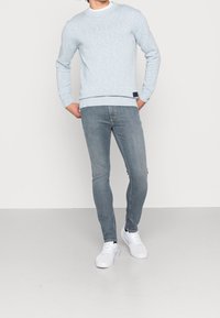 Suéter azul claro con motas, de cuello redondo y mangas largas, combinado con jeans grises ajustados y zapatillas blancas. Detalle de etiqueta negra en el suéter.