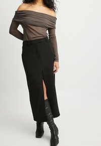 Bruine off-shoulder top met geplooide textuur, gecombineerd met een zwarte lange rok met hoge split en een ceintuur in de taille. Zwarte haklaarzen.