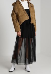 Femme portant une doudoune beige sur une robe maxi noire transparente avec une petite combinaison noire, des bottines blanches et un haut à col roulé blanc.