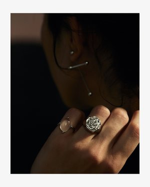 To ringe på en hånd: den ene med et rosemotiv i blankt sølv, og den anden med en oval ædelsten indfattet i sølv, med runde detaljer.