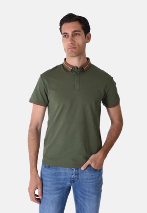Uomo che indossa una polo verde a maniche corte con colletto e bordi delle maniche marroni, abbinata a jeans blu, in piedi con una mano in tasca.