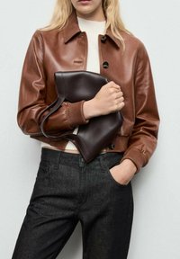 Bruine leren cropped jack met een knoopsluiting, gecombineerd met donkerdenim broek. Houdt een donkerbruine tas met een gebogen ontwerp en handvatten vast.