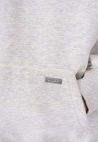 Sv�ětle šedá látka mikiny s klokaní kapsou, která má na přední straně malou obdélníkovou kovovou značkovou visačku.