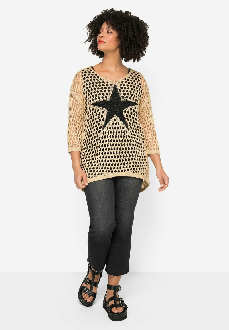 Beige háčkovaný top se velkým černým hvězdným vzorem, tříčtvrteční rukávy a oversized střih, spárovaný s tmavými zkrácenými džíny a černými sandály.
