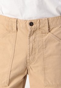 Beige-cotton-housut, joissa on vetoketju ja yksi ruskea nappi, kulmikkaat etutaskut ja teksturoitu pinta.