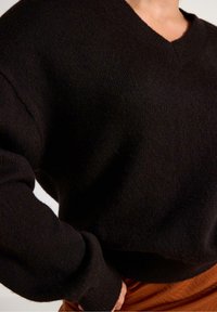 Detailní záběr na osobu, která má na sobě černý pletený svetr s V-výstřihem a dlouhými rukávy, a hnědé kalhoty, přičemž ruka spočívá na bocích.