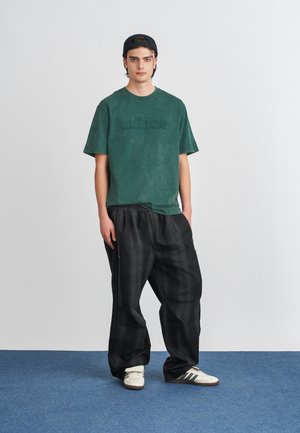 Zaļš kokvilnas T-krekls ar izspiestu "adidas" logotipu, savienots ar brīvām melnām svītru biksēm un krēmkrāsas kedām ar melnām svītrām.