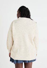 Beige geribbelde trui met een hoge kraag, oversized vorm en verlaagde schouders, gedragen over een denimrok. Heeft een gestructureerde stof.