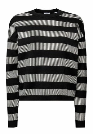 Personne aux longs cheveux bruns portant un pull rayé noir et blanc et un pantalon noir, tirant sur l'ourlet du pull, montrant des bagues en or et un bracelet.