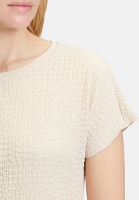 Koszulka z krótkim rękawem w fakturowanym beżowym materiale o miękkiej, pomarszczonej powierzchni i okrągłym dekoltem z kontrastowym wykończeniem.