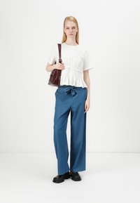 Haut blanc à manches courtes avec une texture froncée, associé à un pantalon en denim bleu ample et des chaussures noires épaisses. Le mannequin porte un sac bourgogne.