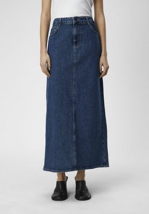 Femme portant une longue jupe droite en denim bleu foncé, un top blanc sans manches et des chaussures noires sans lacets, debout sur un fond gris.