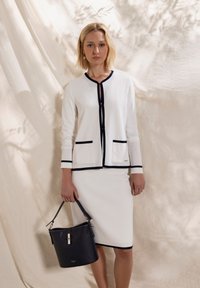 Witte cardigan met marineblauwe bies en zakken, gecombineerd met een witte rok. Houdt een zwarte leren handtas met zilveren accenten vast.