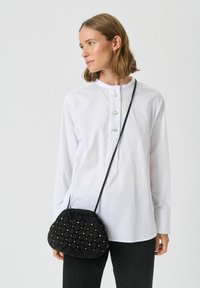 Must karvane kott ümarate kujutistega, millel on dekoratiivsed nupud, kantakse peene rihmaga. Mudel kannab valget särki ja musti pükse.