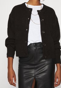 Tmavě hnědý pletený kardigan s kulatým výstřihem, zapínáním na knoflíky a žebrovanou strukturou; nošený přes bílé tričko a lesklou černou sukni.